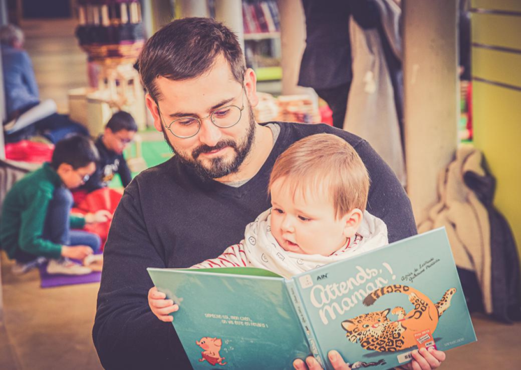 Un petit enfant est installé sur les genoux d'un homme et lit un album dont le titre est attends maman 