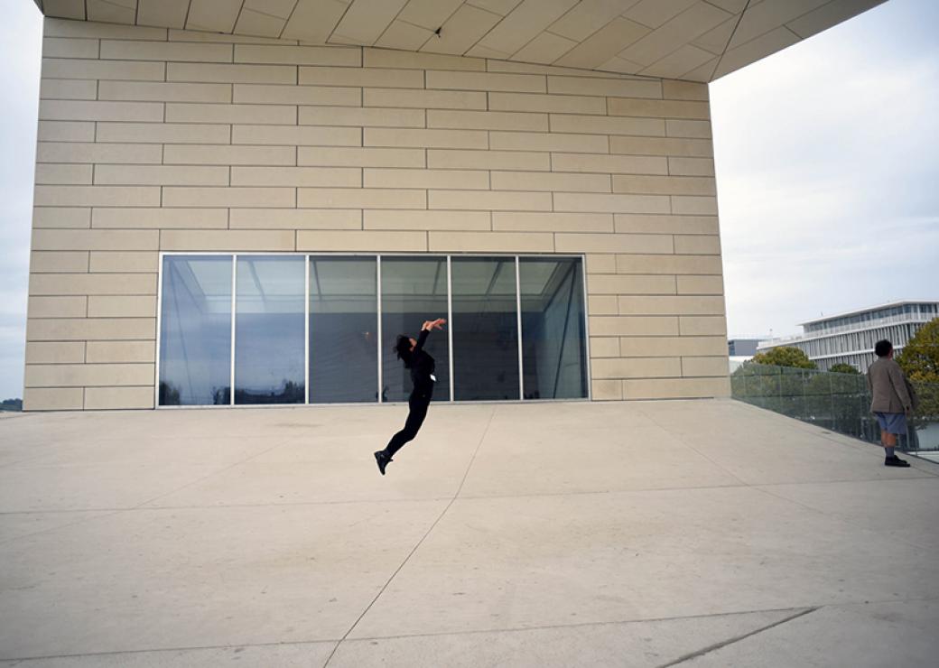 Sur le parvis d'un bâtiment moderne, une personne s'élance dans un saut les bras vers le ciel