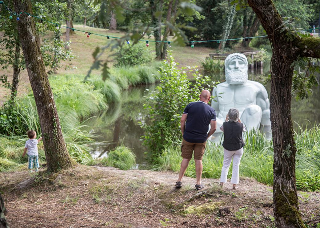 Au milieu d'un étang, une statue d'un dieu grec est installée, le bas du corps immergé dans l'eau, comme pour un bain. Depuis la berge, un couple et un enfant regardent la statue