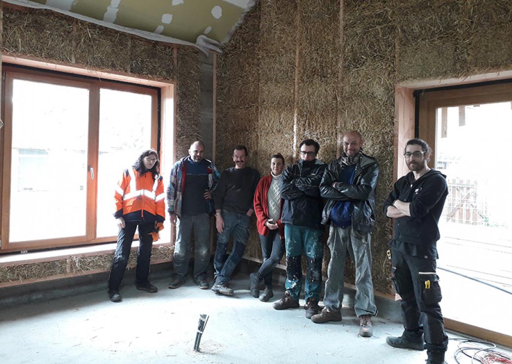 A l'intérieur d'un bâtiment dont les murs sont en paille, 7 personnes posent devant l'objectif du photographe