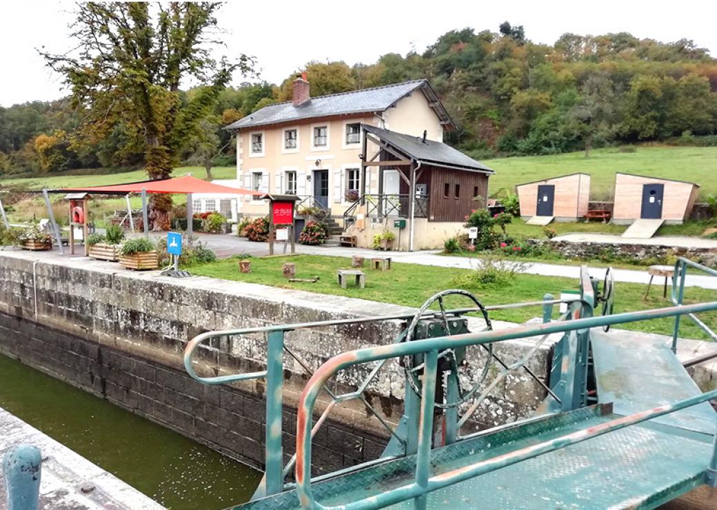 Photo de la maison de l'éclusier, prise depuis l'écluse. À droite de la maison, deux petites cabanes de bois.