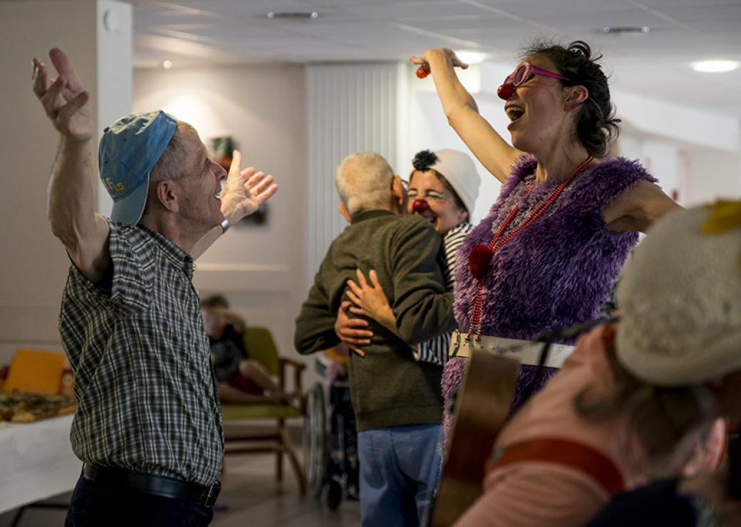 Une femme clown ouvre les bras à un homme agé qui ouvre lui aussi ses bras. En arrière-plan, une femme clown étreint un homme aux cheveux blancs.