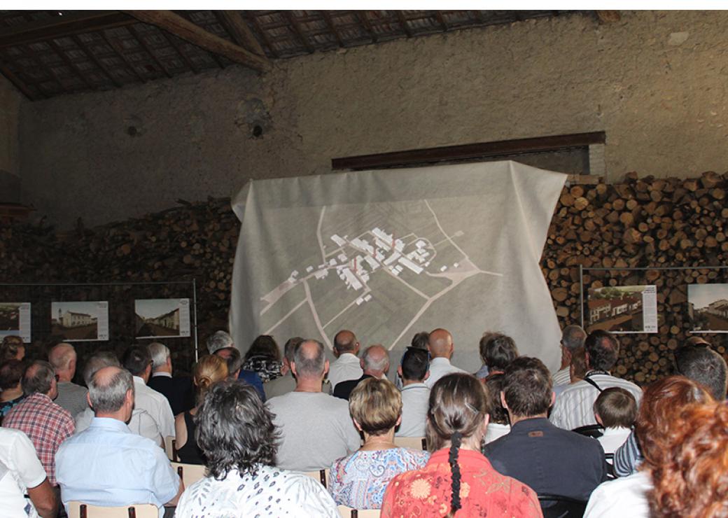 Dans une grange, un drap est tendu contre des rondins de bois. Une photo aérienne est projetée sur ce drap, face à une trentaine de personnes assises