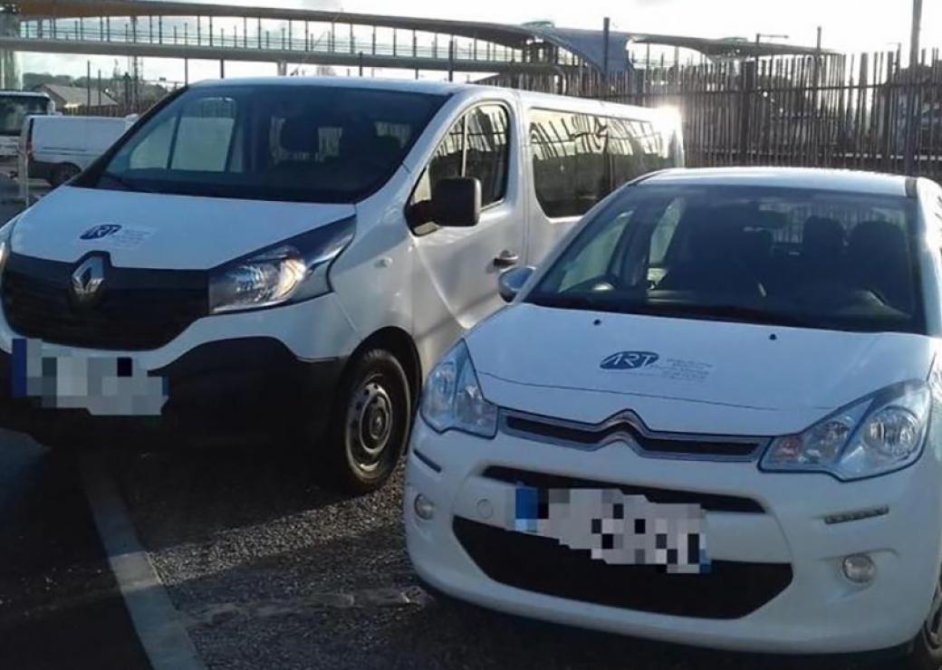 Deux véhicules blancs, un utilitaire et une petite voiture, garés sur le parking devant la passerelle d'une gare