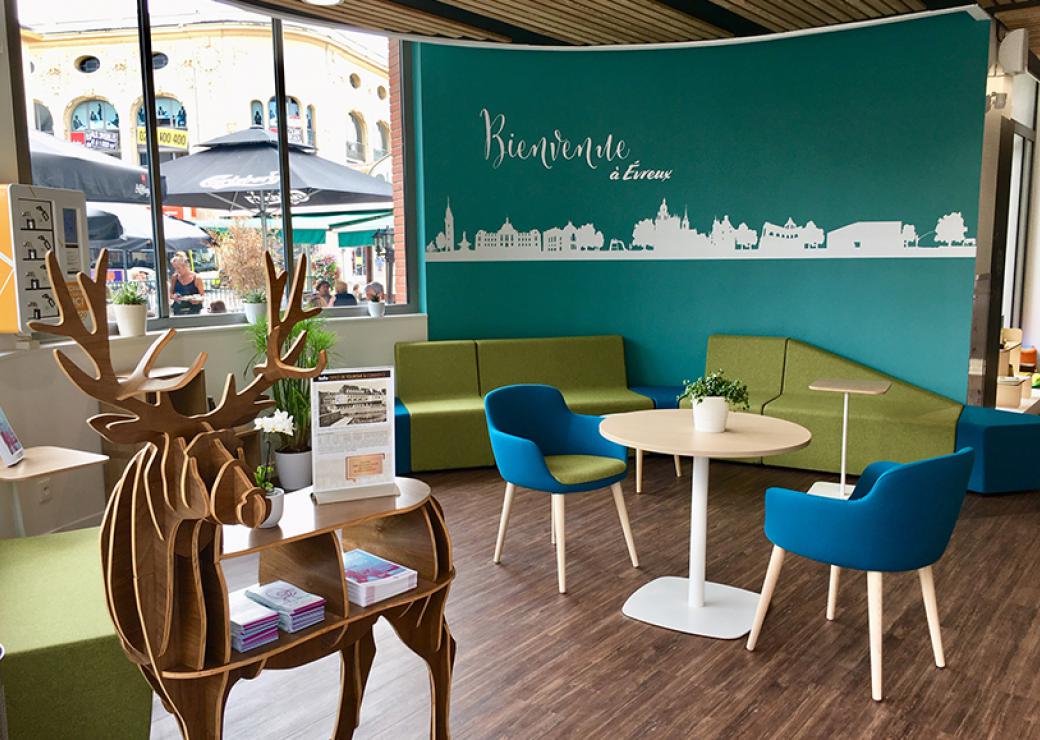 Vue d'un salon d'accueil. Sur le mur du fond, on peut lire "Bienvenue à Evreux". Par la fenêtre, on distingue des parasols abritant des terrasses de café