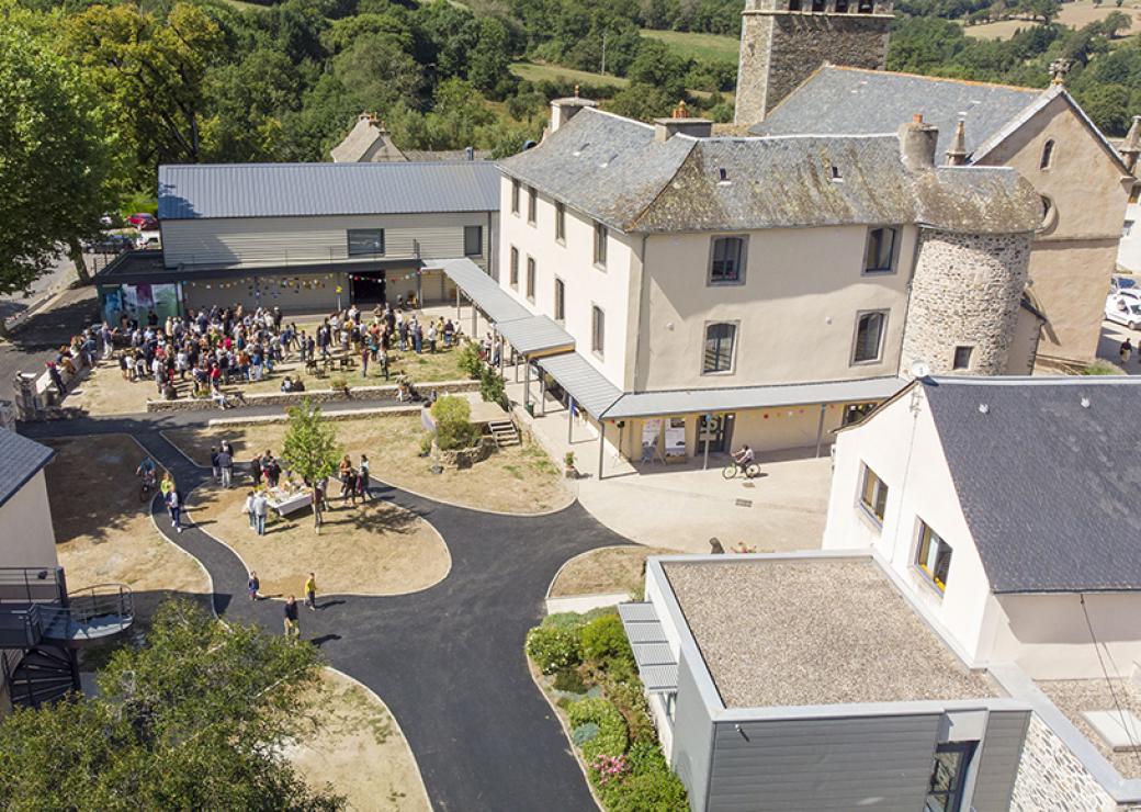 Vue aérienne d'un cœur de village où se mêlent constructions médiévales et bâtiments modernes. Sur la place, on distingue les silhouettes d'une soixantaine de personnes