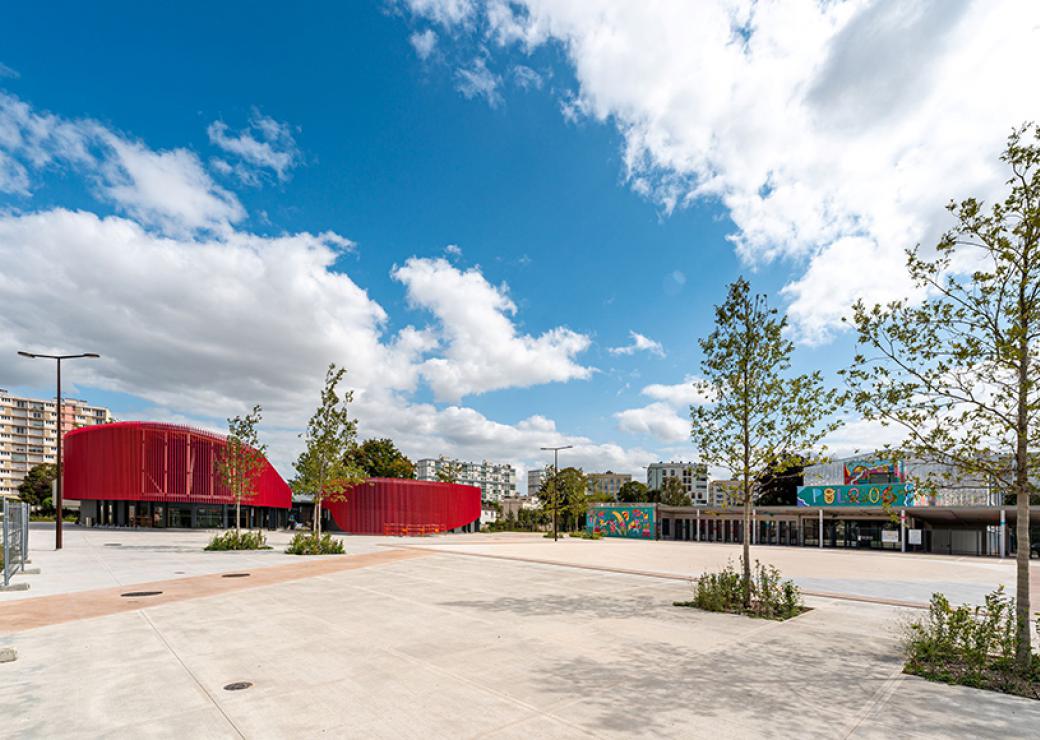 Une large place dallée plantée de quatre jeunes arbres est bordée par un bâtiment moderne rouge et au loin des immeubles d'habitation