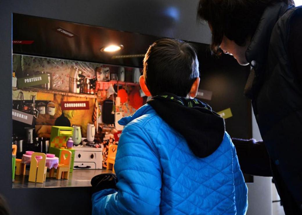 Un enfant et un adulte, tous deux dos au photographe, regardent le décor d'une cuisine en miniature 