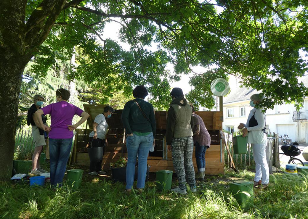 Sous les branches d'un arbre aux feuilles très vertes, des personnes se tiennent devant de gros bacs de bois. Certaines tiennent un seau vert à la main, d'autres l'ont à leurs pieds