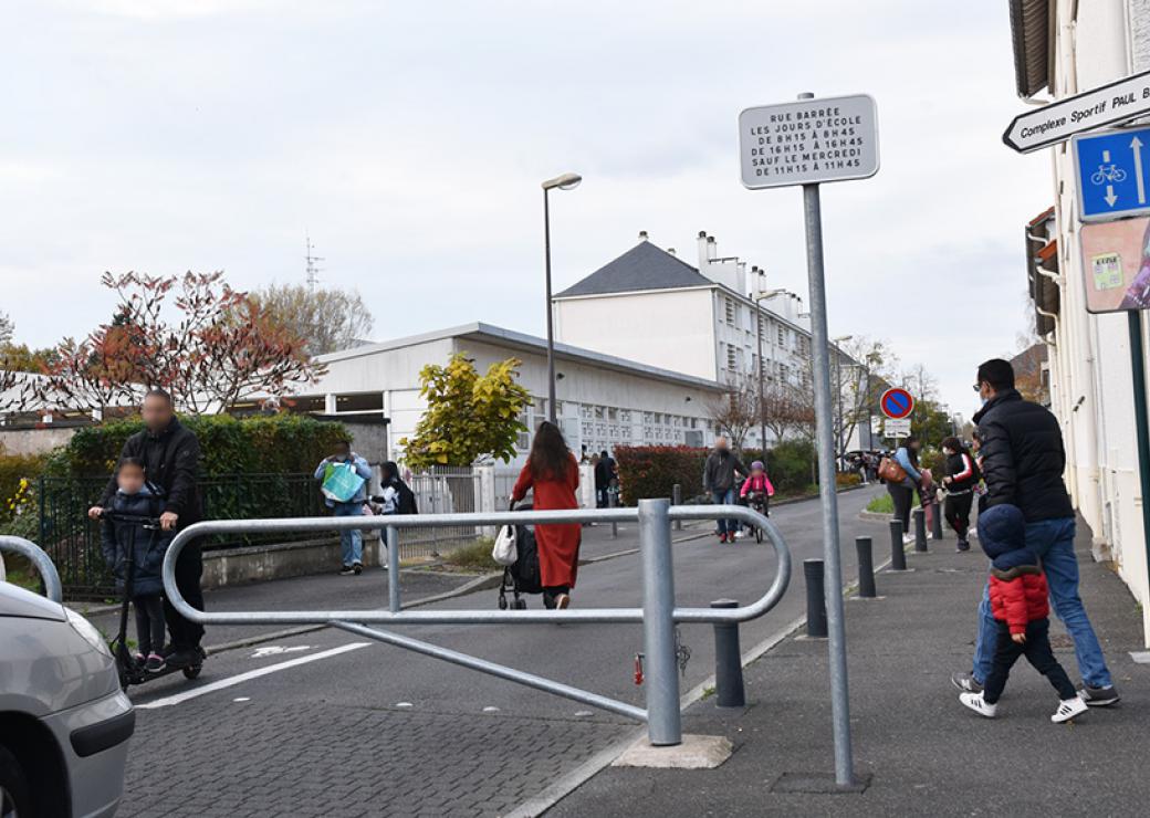 Vue d'une rue fermée par une barrière métallique, où des adultes marchent à côté d'enfants