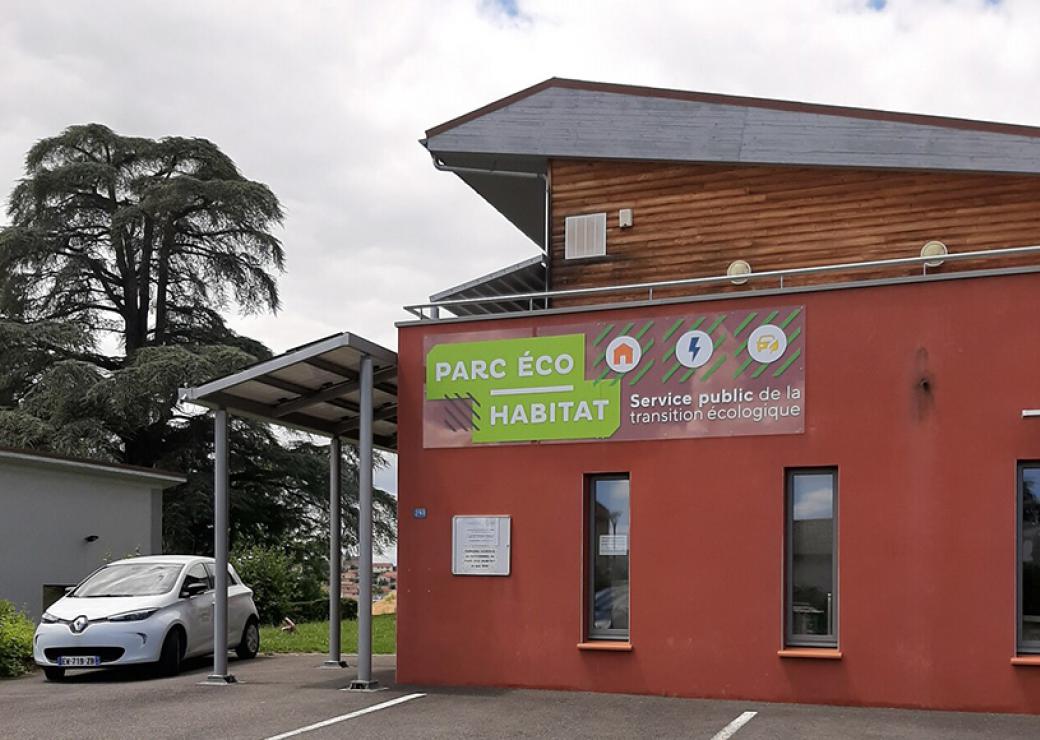 Vue large d'un bâtiment moderne, enduis rouge et bardage de bois. Sur une enseigne on peut lire "Parc éco habitat, service public de la transition écologique" 