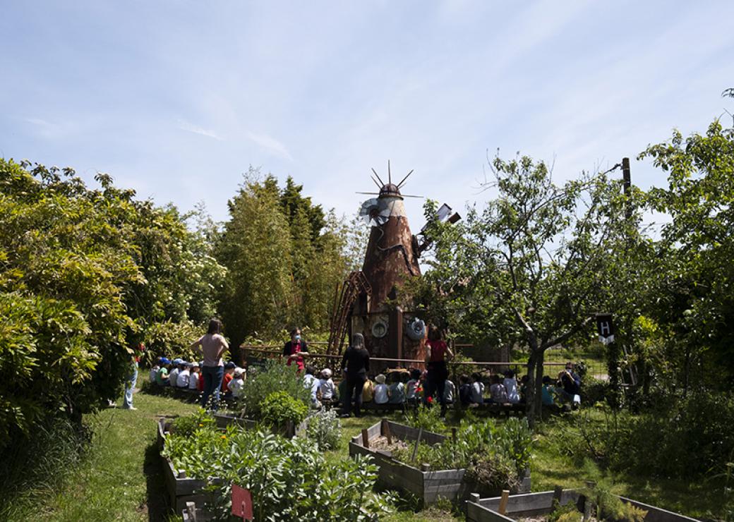 Dans un jardin sous le ciel bleu, des enfants sont assis sur des bancs, autour d'une grande sculpture de bois