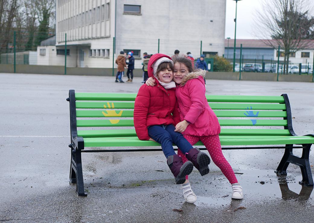 Une enfant se serre contre une autre enfant, elles sourient, assises sur un banc vert