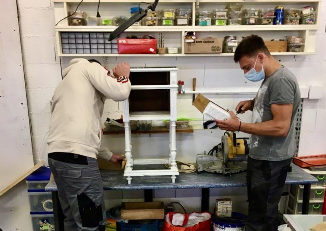 Dans un atelier, deux jeunes personnes réparent une table de nuit