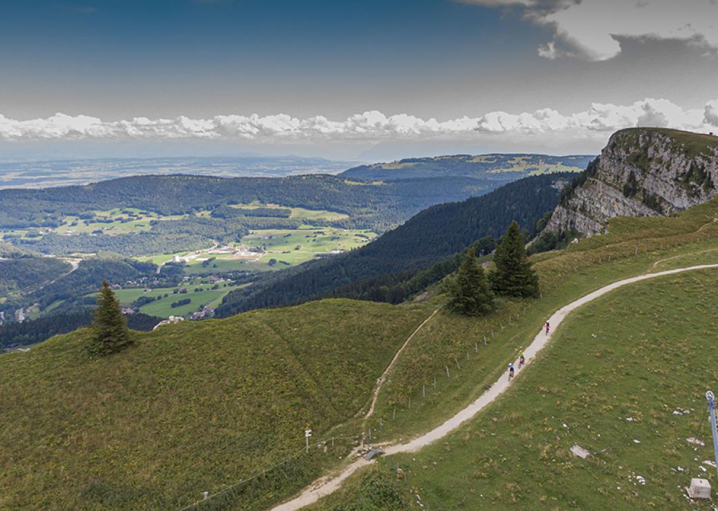 Vue panoramique d'un paysage boisé et vallonné. Au premier plan, sur la crête d'une colline, un chemin de randonnée, on distingue 3 silhouettes