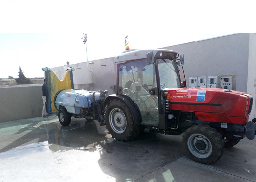 Un tracteur rouge équipé d'une remorque est stationné sur une plateforme de béton. On voit une flaque d'eau au sol.