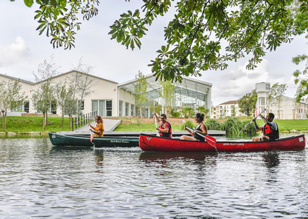 Deux canoés, avec chacun deux rameurs, naviguent sur une pièce d'eau qui longe un pelouse et des bâtiments modernes