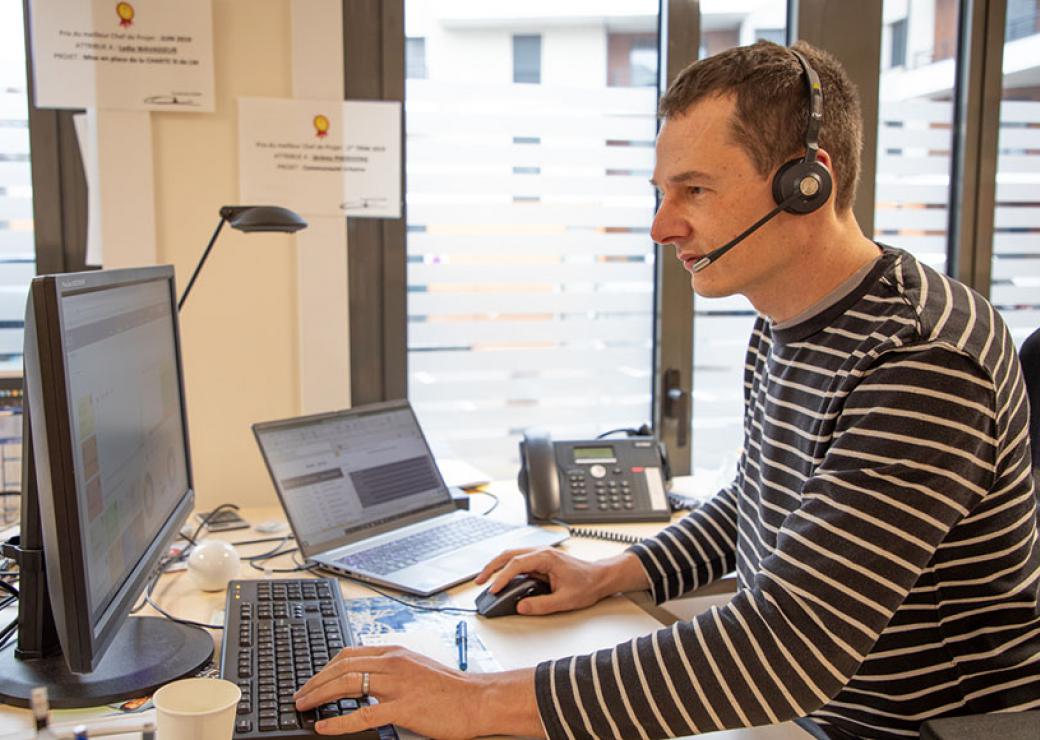 Plan moyen d'un homme équipé d'un micro et d'un casque audio, de profil. Il est face à deux écrans d'ordinateur