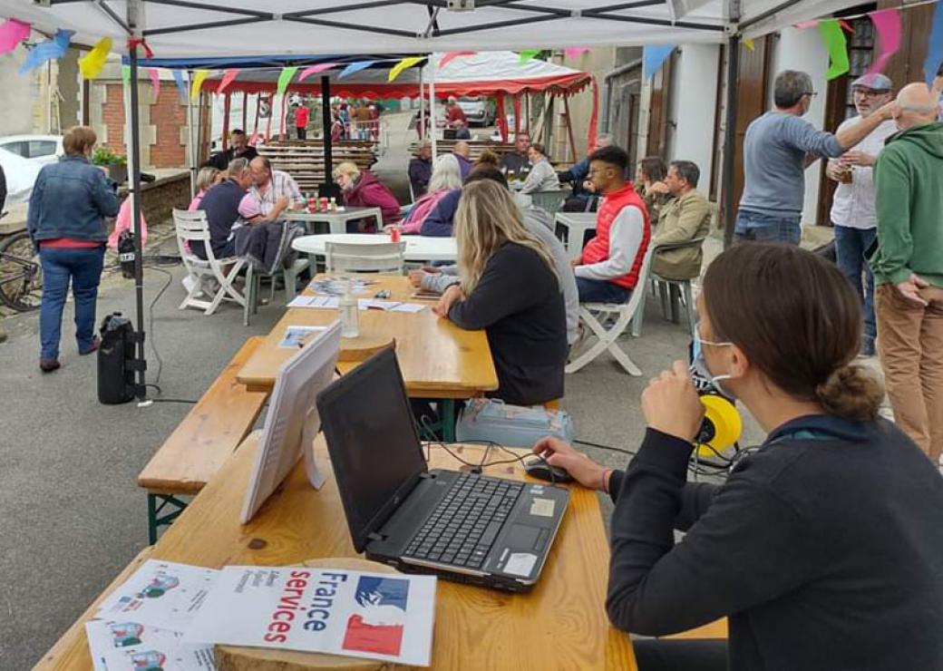 Dans la rue, sous des parapluie de forain, des tables sont installées. On voit des personnes assises qui discutent. Au premier plan, une jeune fille fait face à un ordinateur. Un feuille indique France service, avec les couleurs bleu blanc rouge