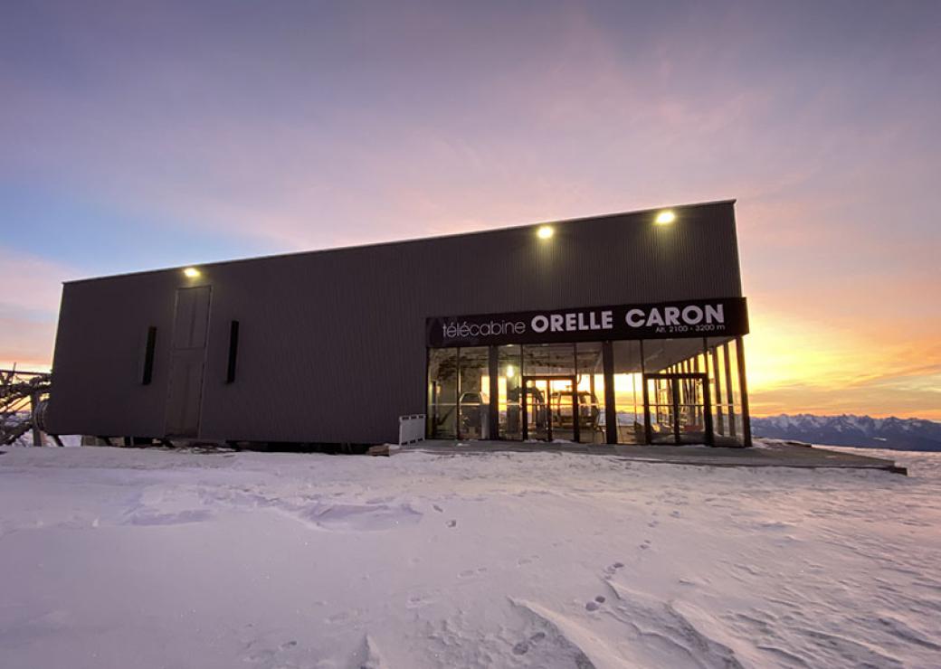 Vue d'un grand bâtiment posé sur la neige, avec le soleil rasant. On peut lire télécabine Orelle Caron