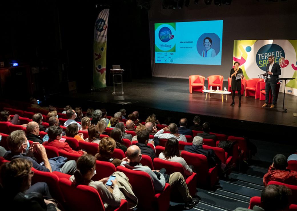 Dans une salle de spectacle plein de spectateurs, deux personnes sont sur scène. Derrière elles, on peut lire "terre de santé" 