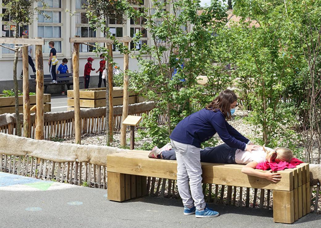 Sur un banc une petite fille est allongée, une autre lui touche le dos. Au fond, un groupe d'enfants. En arrière plan, des buissons