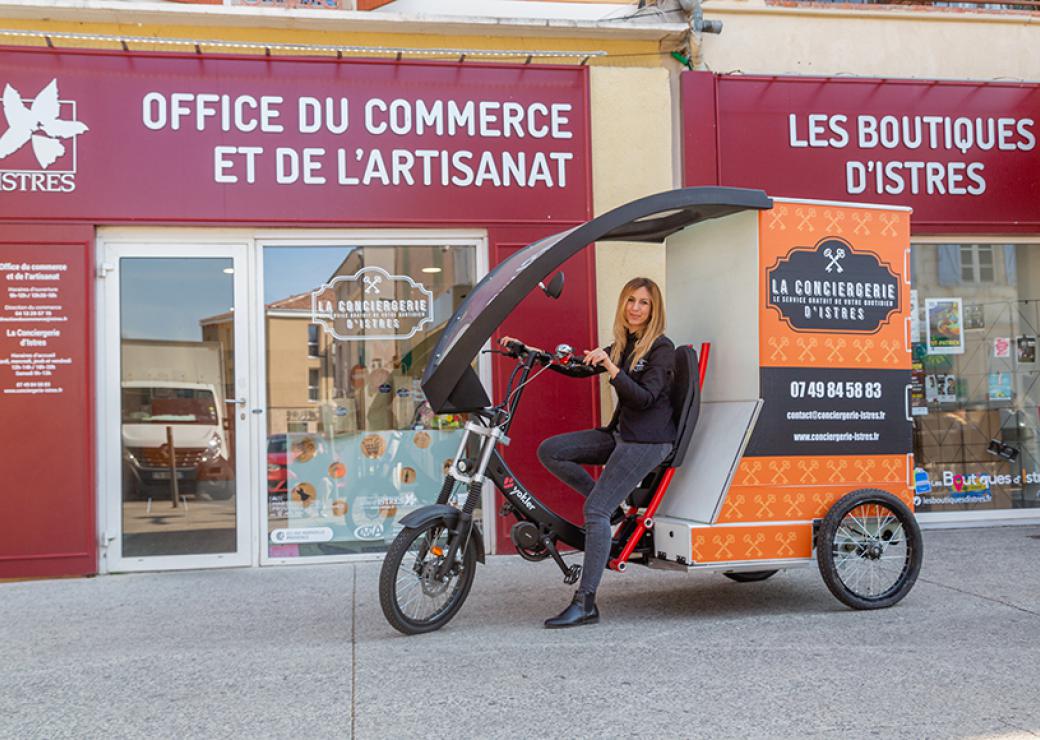 Une jeune femme au guidon d'un triporteur pose devant la devanture d'une boutique où l'on peut lire "office du tourisme et de l'artisanat, les boutiques d'Istres"