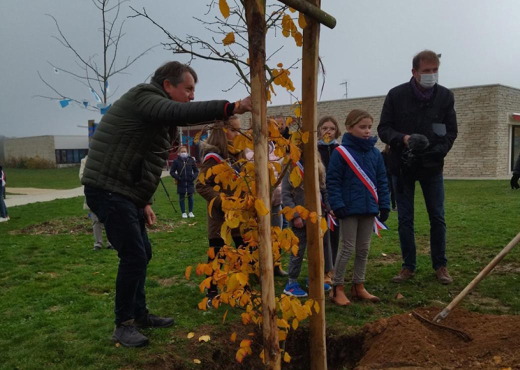 Un homme plante un arbre. A ses côtés, des enfants ceints d'une écharpe tricolore.