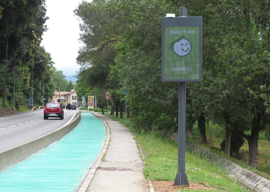 Une petite voiture rouge sur une route, à droite, un panneau numérique avec un écran vert où l'on peut lire Qualité air Respirez!