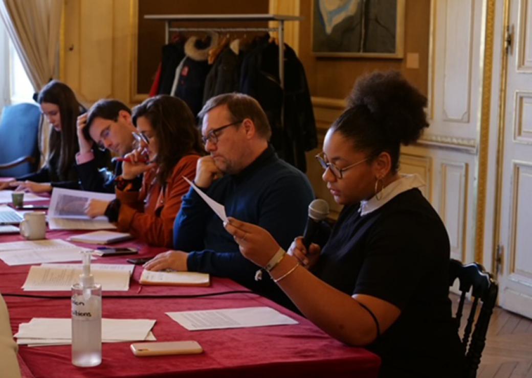 5 personnes assises à une table lisent des documents, l'une des personnes parle avec un micro