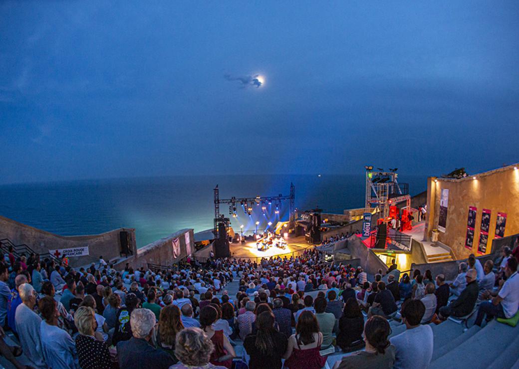 Un public dense est rassemblé dans les gradins face à une scène éclairée par des projecteurs. Derrière la scène, la mer.