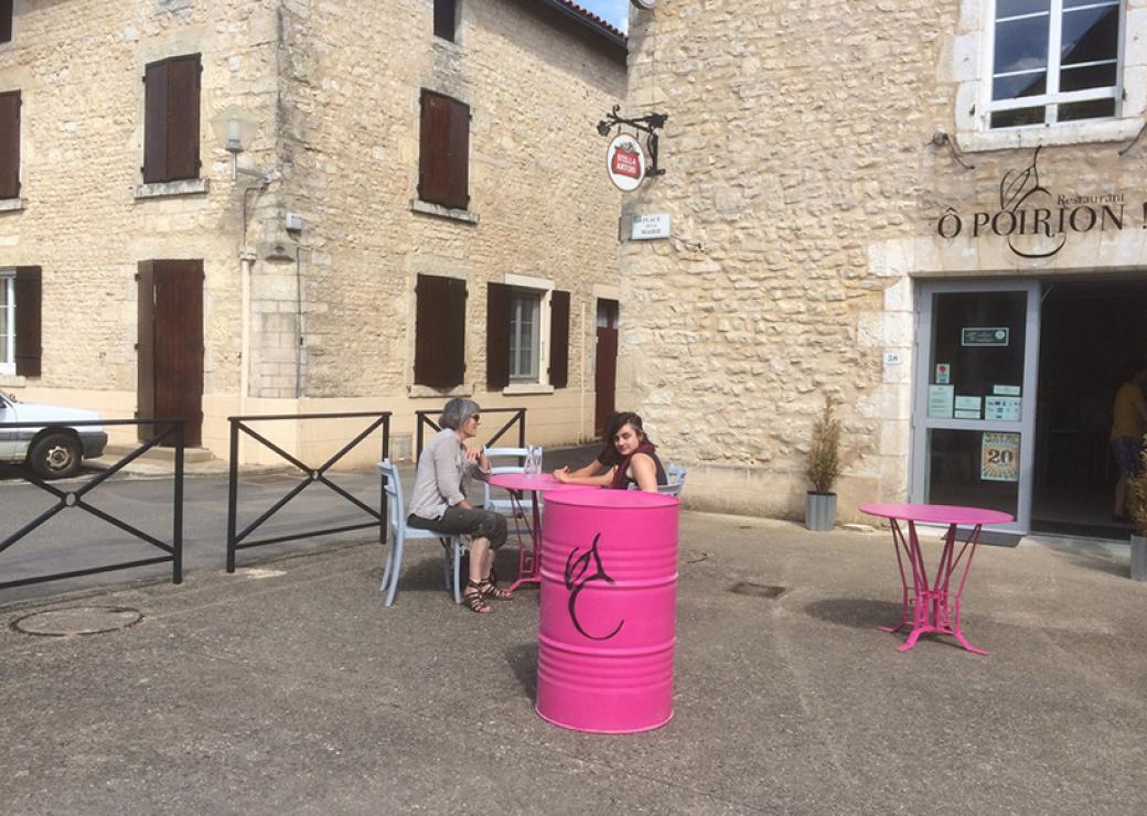 Deux femmes sont attablées à la table d'une terrasse, entourée de bâtiments aux pierres apparentes