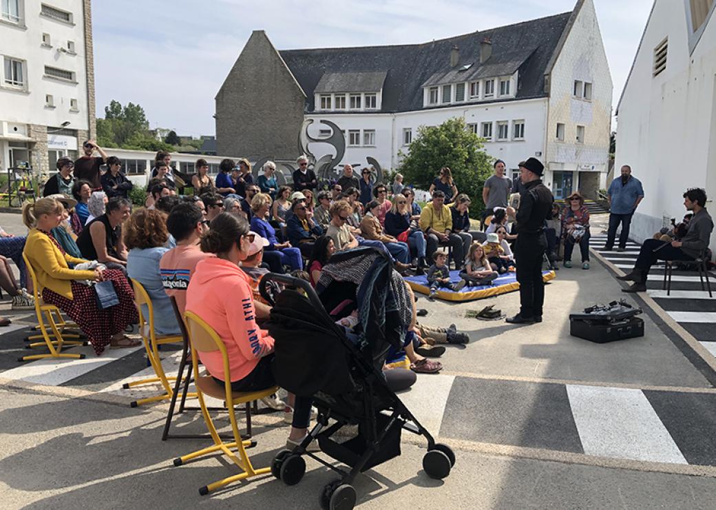 Dans la rue, assises en demi cercle, des personnes assistent à une représentation