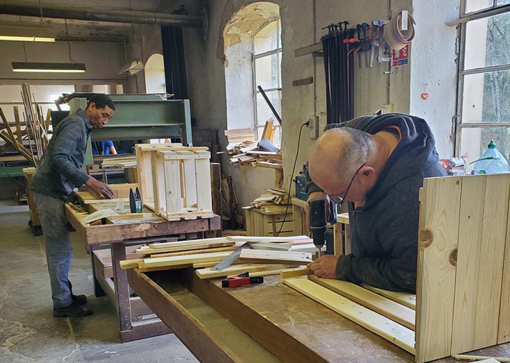 Dans un ateliers, deux hommes sont affairés sur des établis. L'un d'entre eux ajuste une perceuse sur une planche de bois