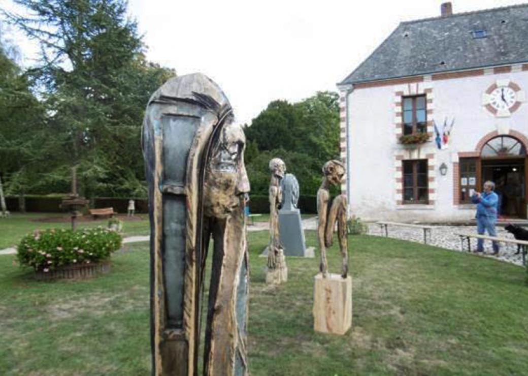 En plein air, de grandes statues de bois sont exposées. A l'arrière plan, un bâtiment orné de drapeaux