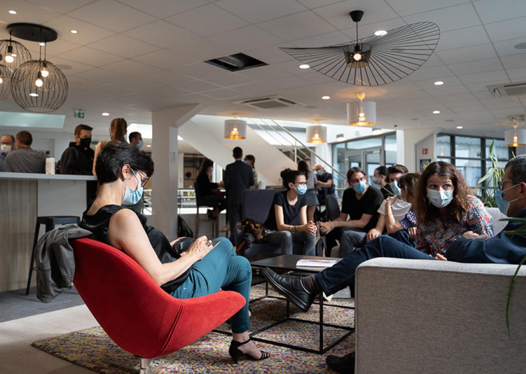 Dans une très grande salle, une quinzaine de personnes portant des masques chirurgicaux sont assises sur des fauteuils bas et des canapés.