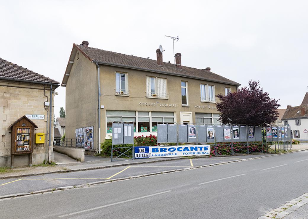 Depuis le bord d'une route, photo d'un bâtiment sur lequel on peut lire groupe scolaire. Une banderole indique aussi Brocante Labbeville Foyer rural 04 juillet  