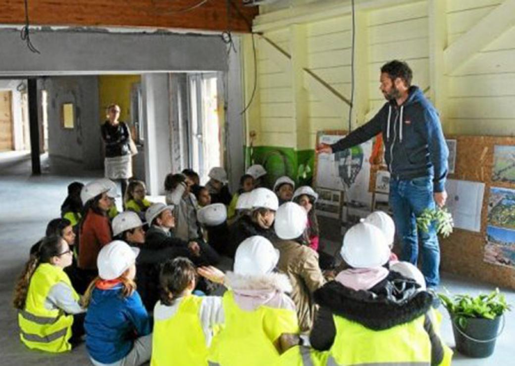 Un groupe d'enfants coiffés de casques de chantier est assis devant un homme qui tient des rameaux dans sa main. Ils sont dans un bâtiment en chantier