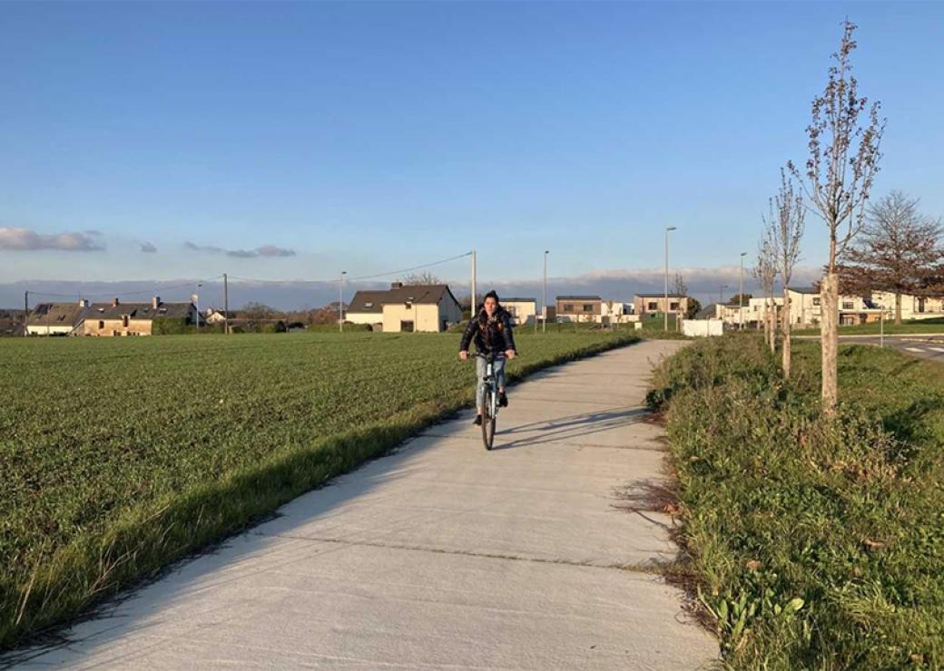 Une personne circule à vélo sur une piste bordée par des champs. Au loin, des pavillons. Ciel bleu