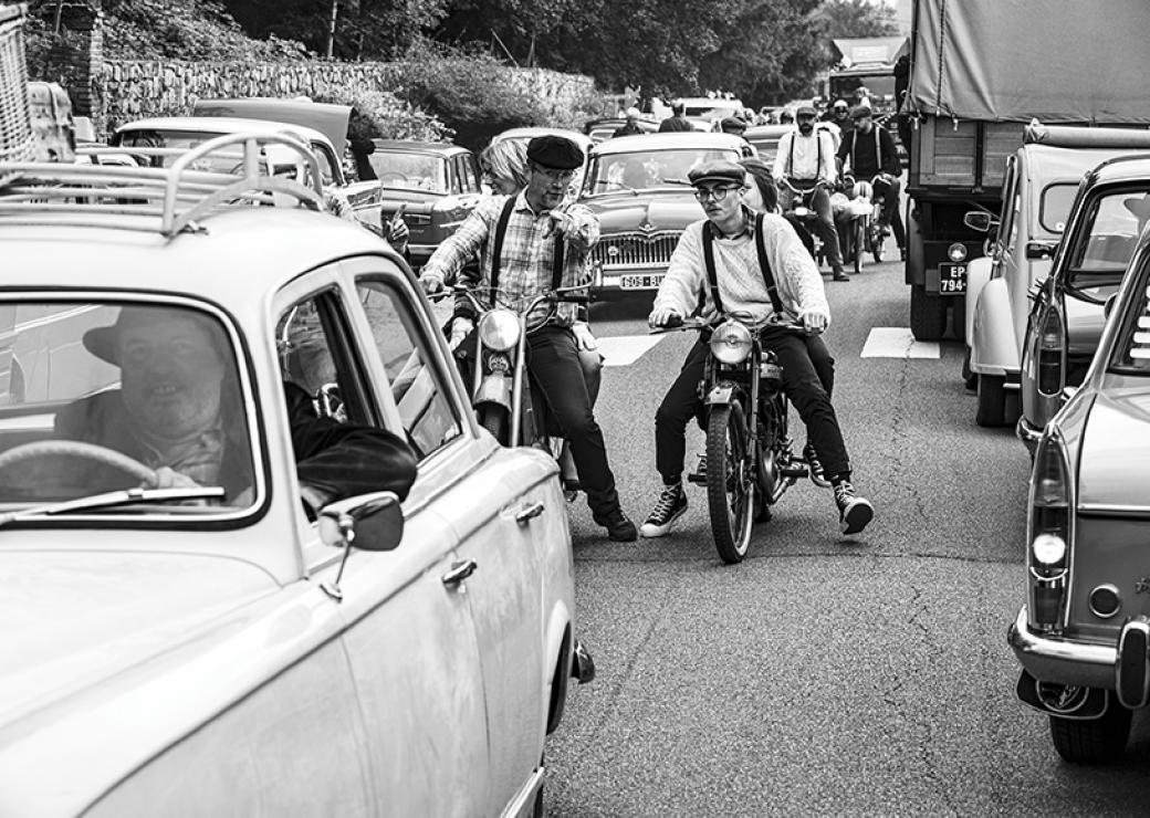 Photo en noir et blanc. Sur une route, un embouteillage de voiture des années cinquante. Au centre, deux motards discutent. Ils portent des bérets et de bretelles