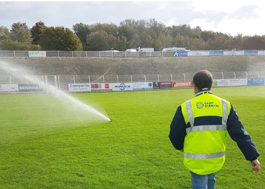 Un homme portant un gilet jaune sur lequel on peut lire "saint-Quentin" marche en direction d'un jet d'eau installé sur une pelouse