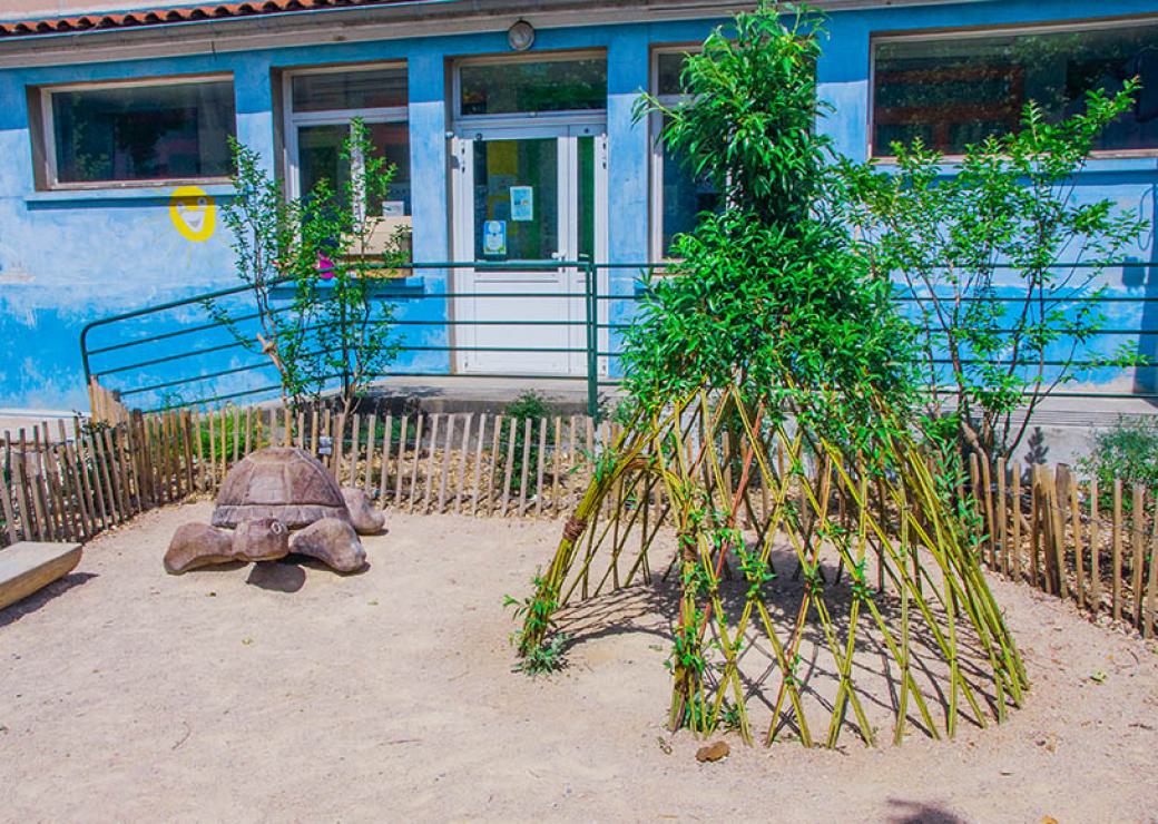 Sur du sable sont posés une tortue en bois et une hutte en roseau. Au fond, un bâtiment bleu