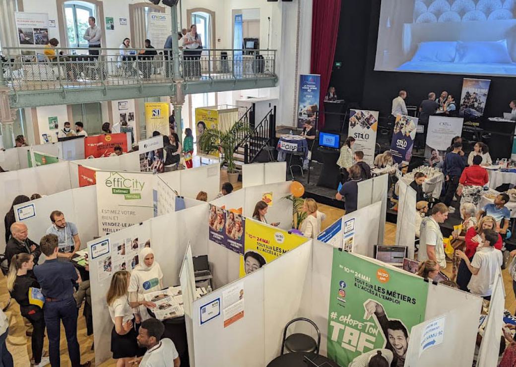 Vue d'en haut, une vaste salle cloisonnée en petits stands. Des personnes évoluent dans ces stands