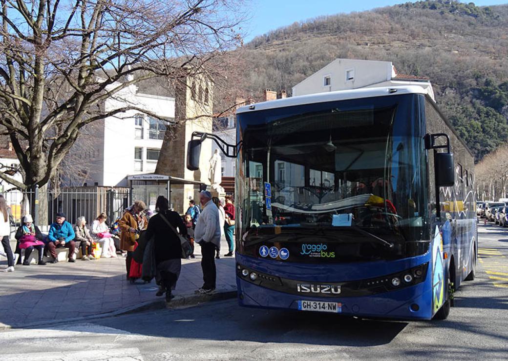 Dans une ville, un autocar est arrêté devant un abribus où sont assises des personnes. En arrière plan, une colline boisée