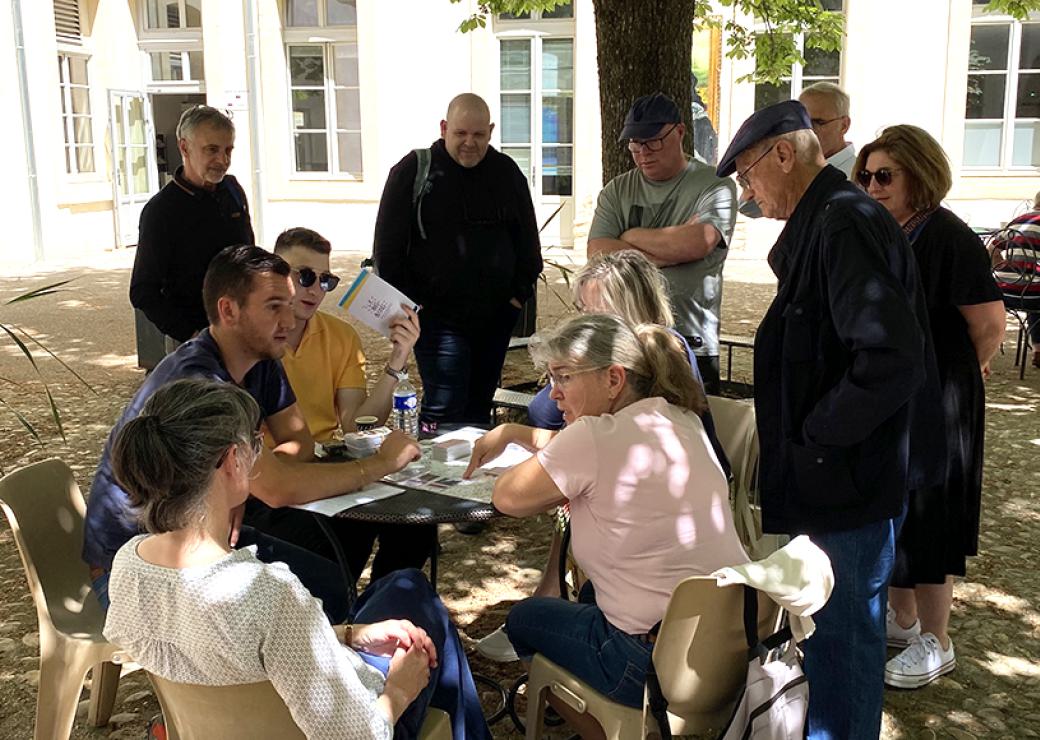 Une dizaine de personnes autour d'une table, certains sont assis, d'autres debout, la table est placée à l'ombre d'un arbre