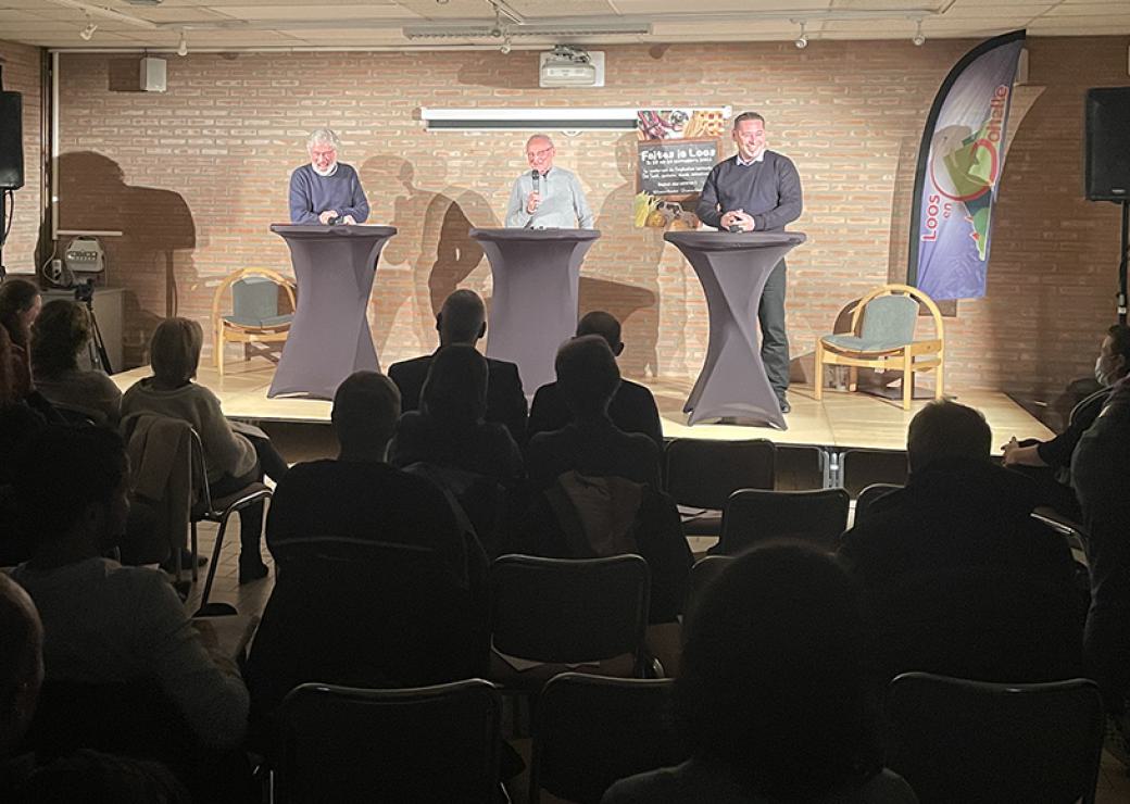 Dans une salle, trois personnes se tiennent sur une estrade devant un public, face à des personnes assises