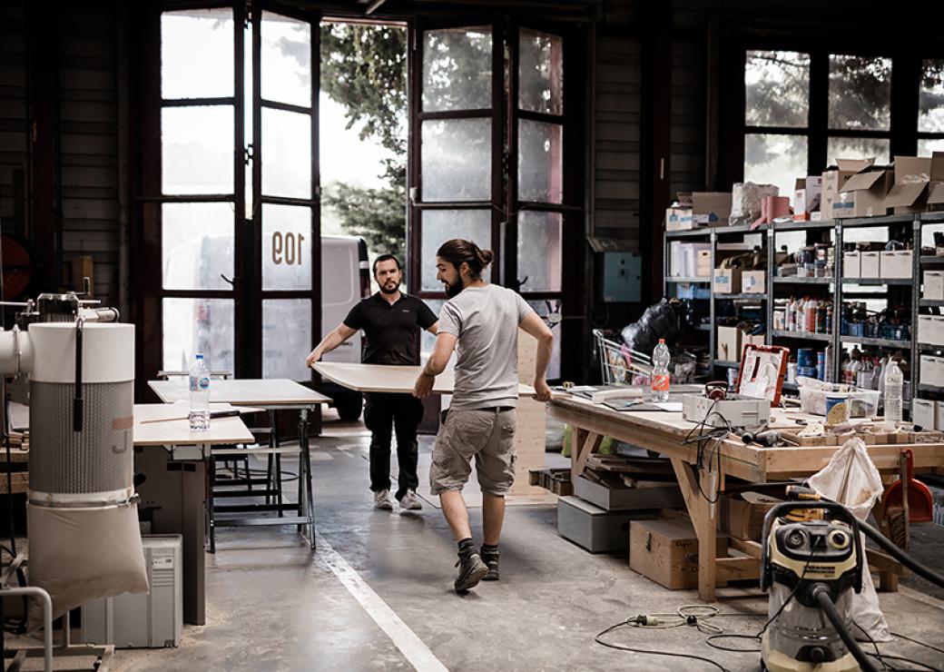 Deux hommes portent un grand panneau de bois à l'intérieur d'un atelier avec de très grandes portes-fenêtres