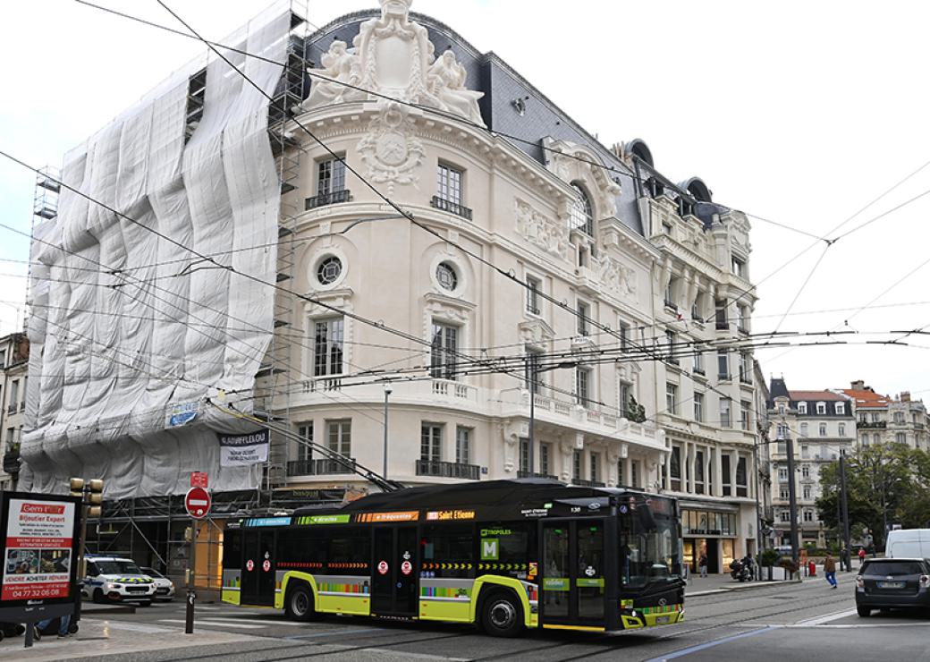 Photo d'un immeuble d'angle aux murs crème et rose poudré, chargés de sculptures et colonnettes. Un voile couvre un échafaudage sur une partie de la façade.