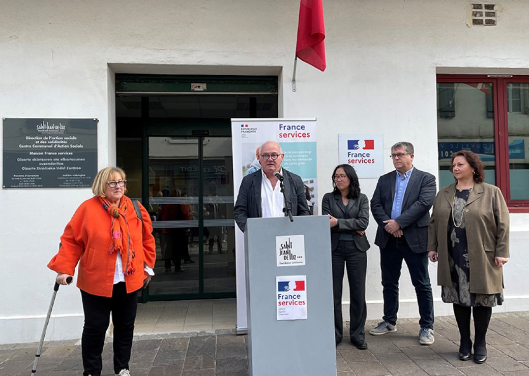 4 personnes encadrent un homme qui parle à un pupitre devant la façade d'un bâtiment. En plusieurs endroit, on peut lire "France services"