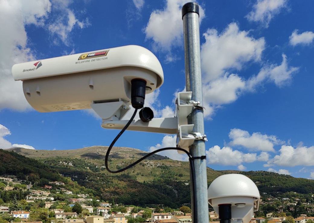 Dans un paysage de collines boisées, un mat équipé de caméras surplombe un village